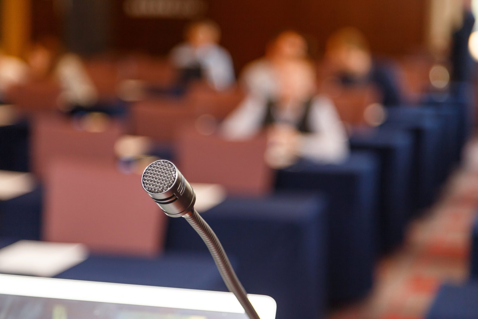 A gooseneck microphone on a desk in a meeting hall, with seated attendees out of focus in rows behind it