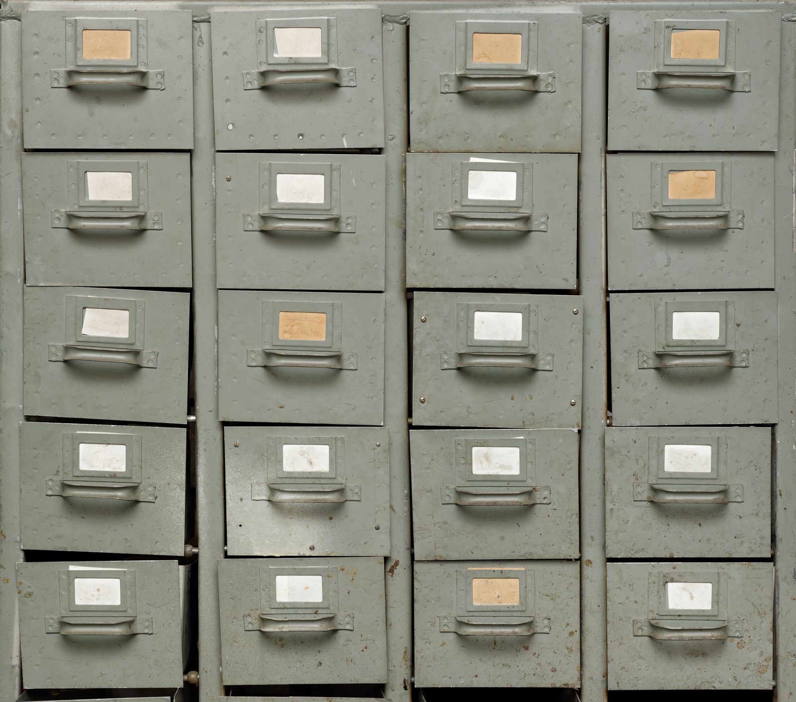 A wall of worn gray metal filing drawers, each with a small paper label holder and a pull handle