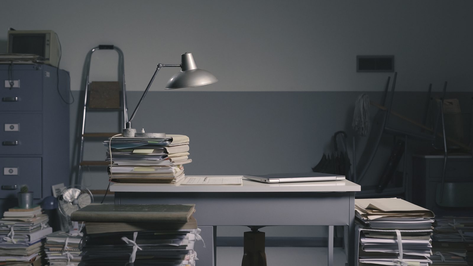 A dim records office with a desk lamp, metal filing cabinets and tall stacks of paper files waiting to be sorted