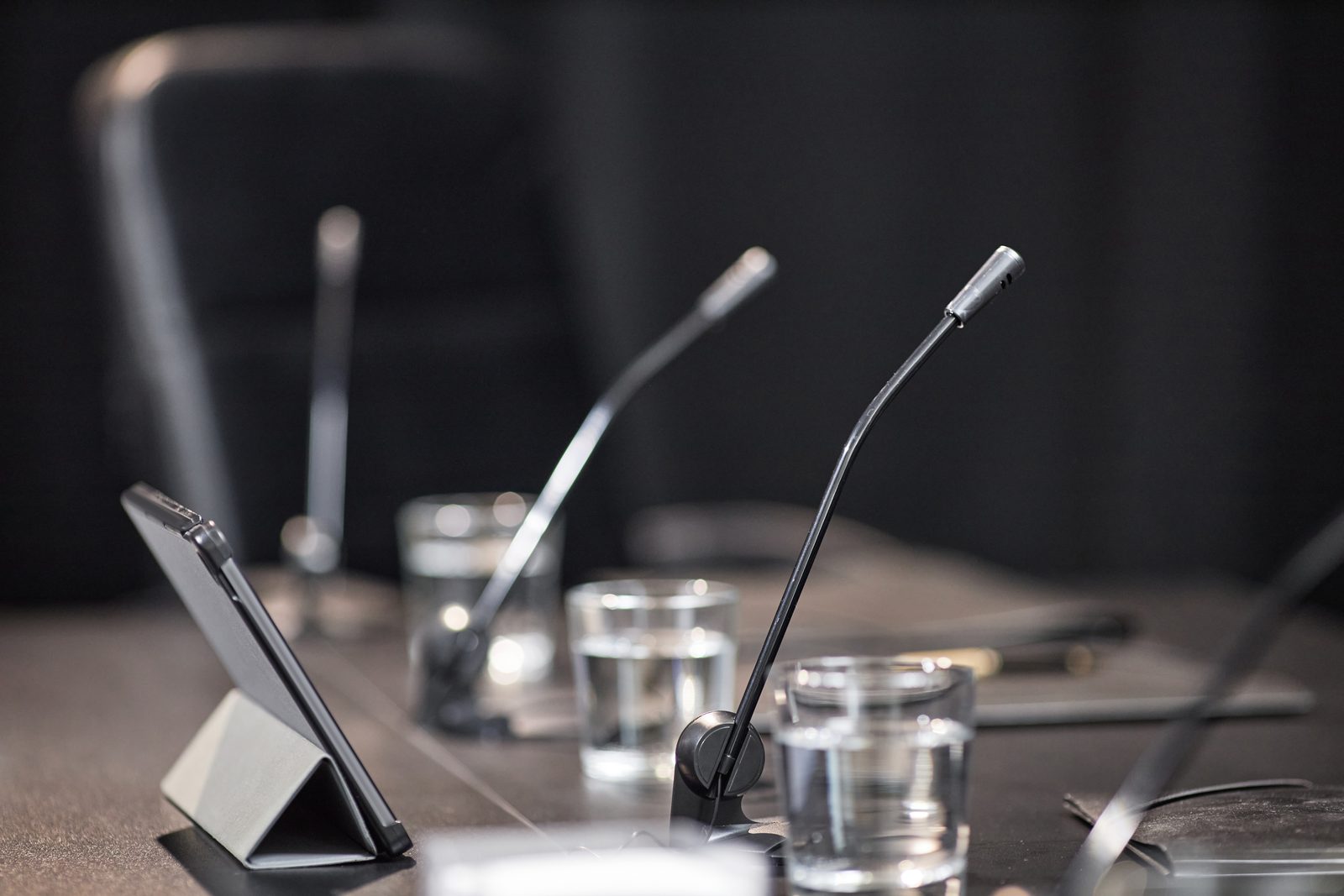 Gooseneck microphones, water glasses and a tablet on a stand sit on a dark table in an empty local government meeting room