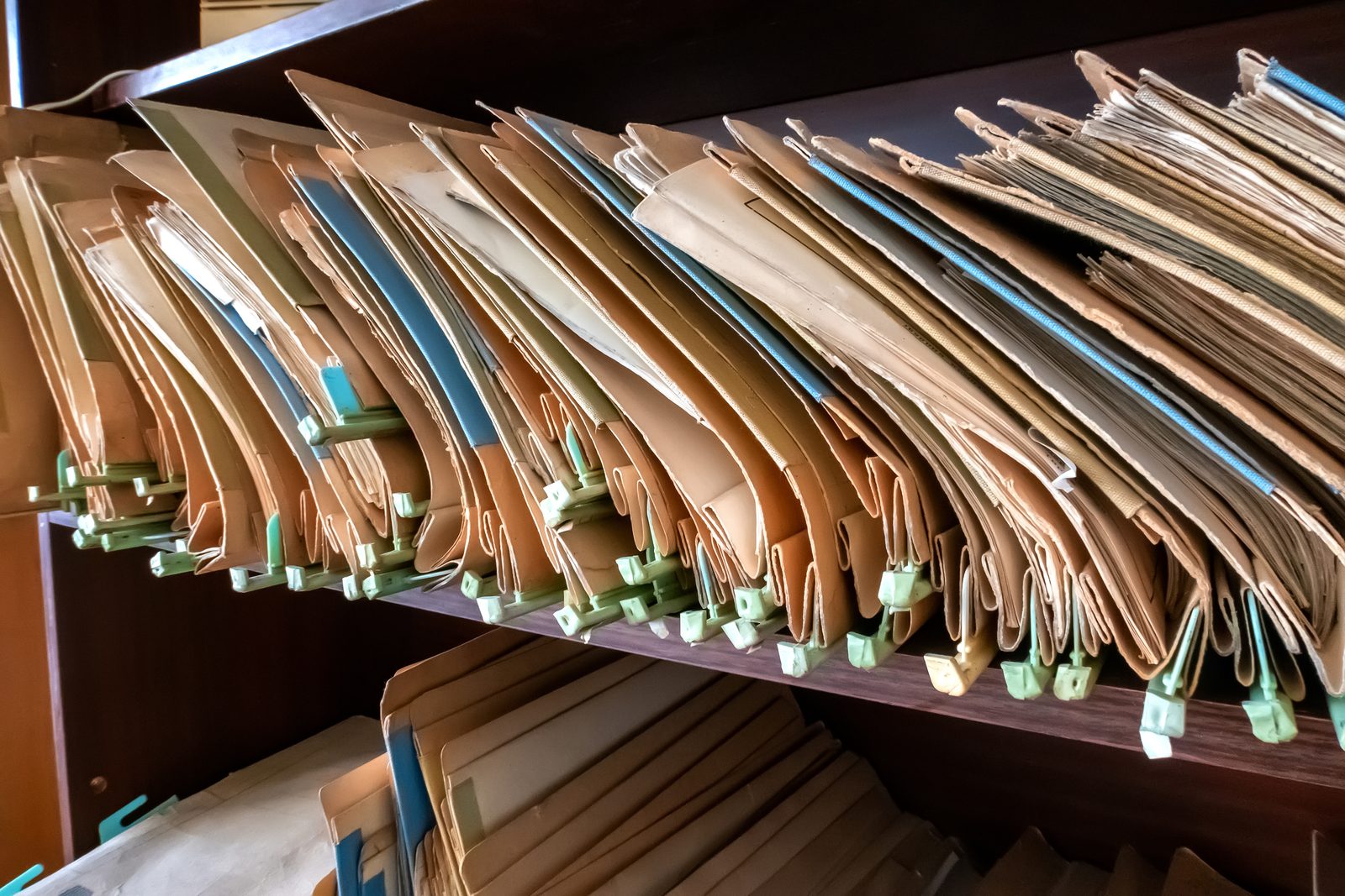 Worn manila file folders with green plastic fasteners packed tightly on a dark wooden records shelf