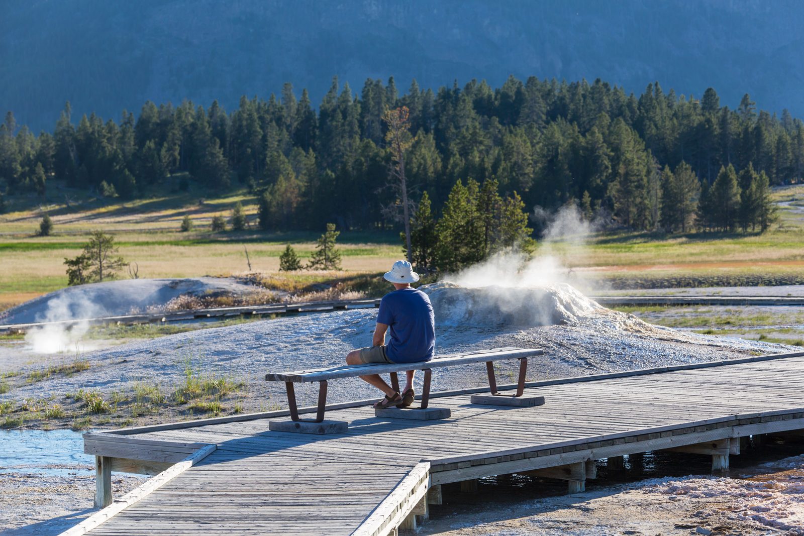 Visitor in a white hat and blue shirt sits on a bench along a wooden boardwalk as steam rises from a geyser basin behind him