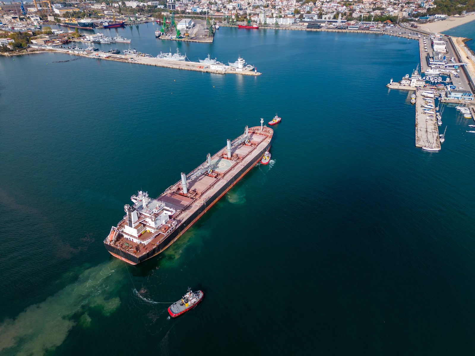 Aerial view of a large bulk carrier leaving a harbor with tugboats alongside, piers and moored vessels lining the shore behind it.