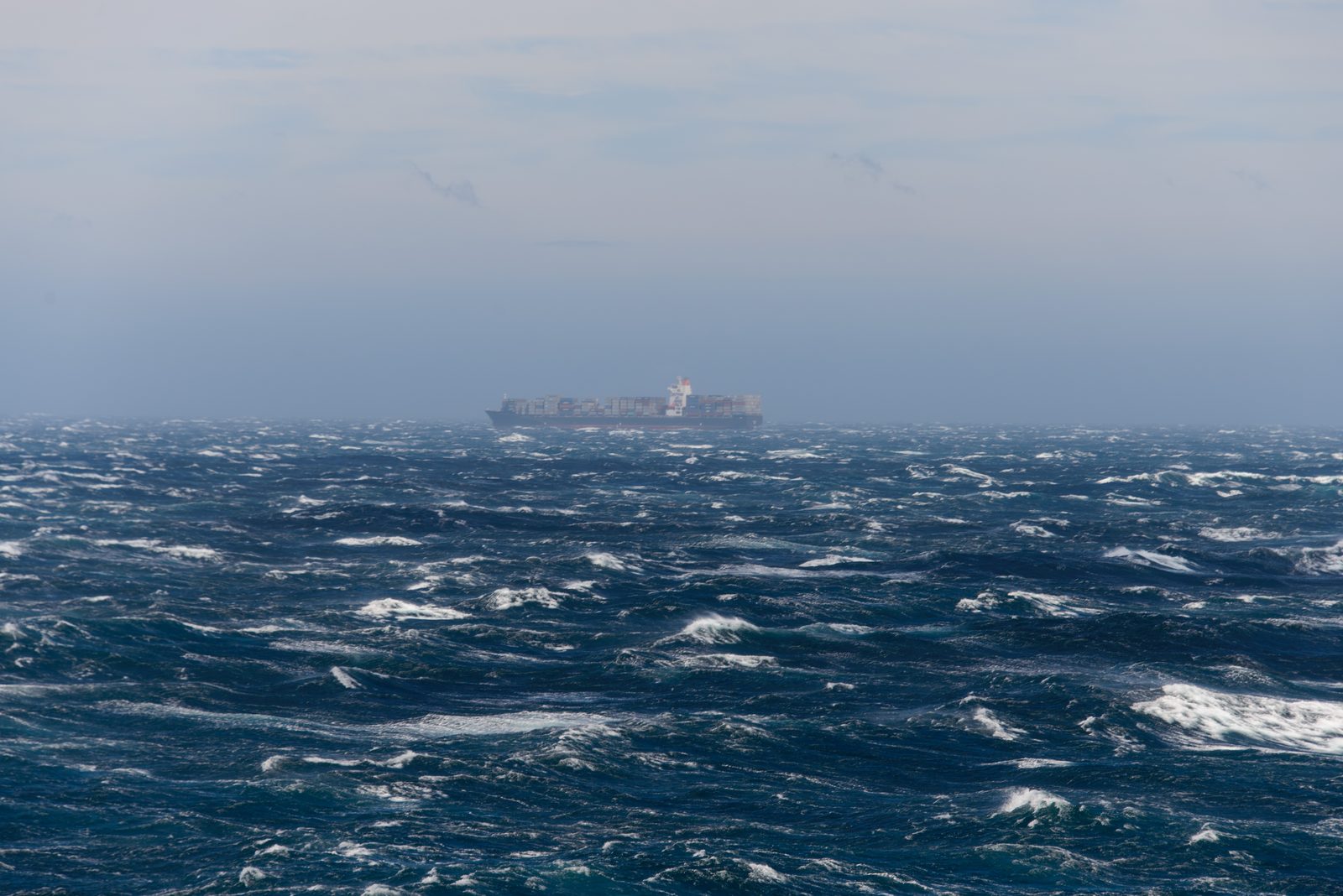 A loaded container ship sits low on the horizon in haze while wind-whipped waves break across the open sea in front of it.