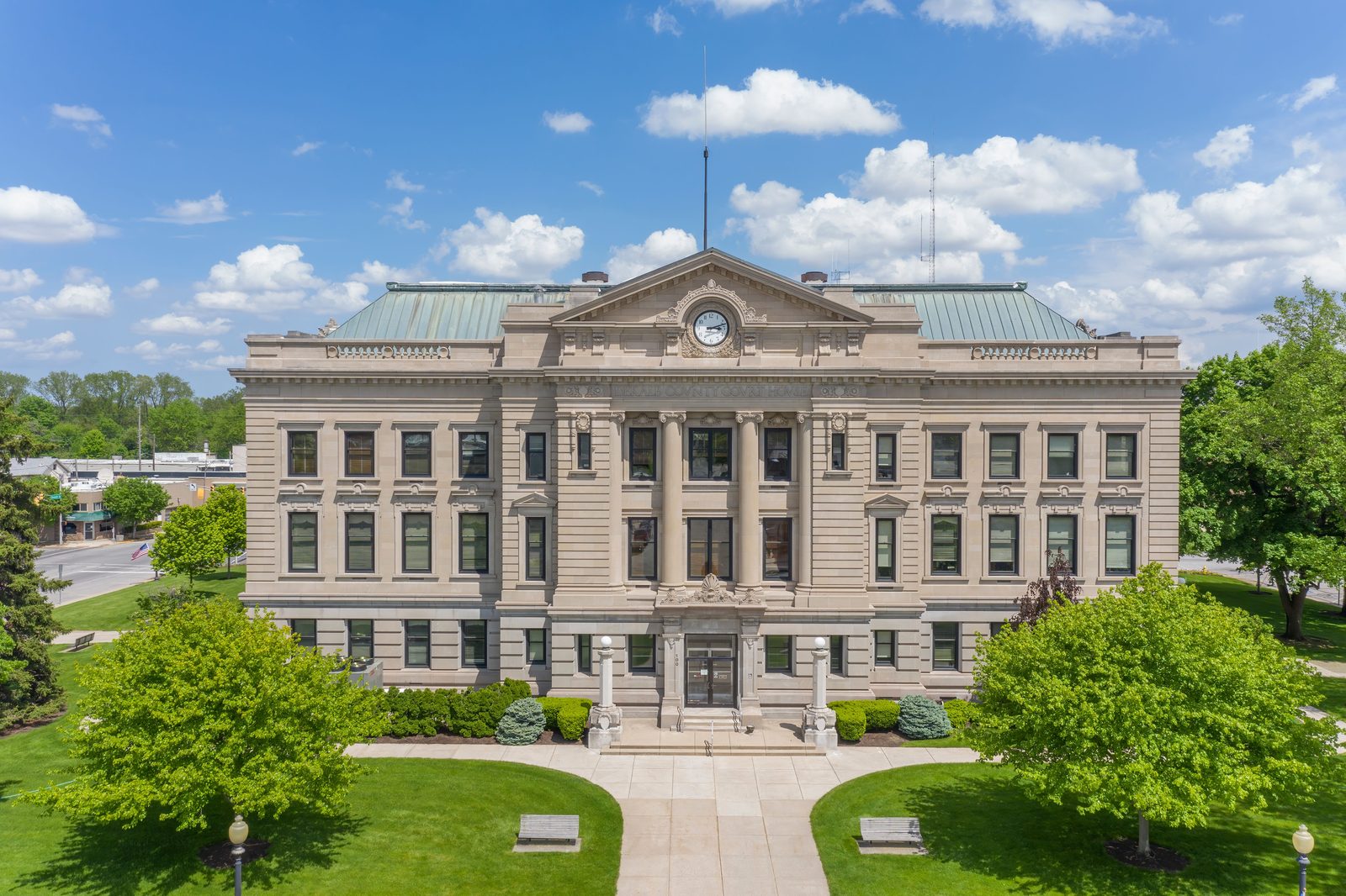 Neoclassical stone county courthouse with a clock in its pediment and a green roof, set behind lawns and trees under a blue sky.