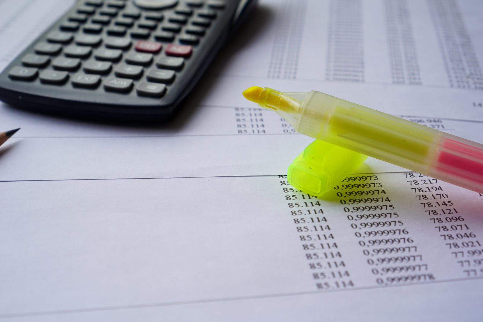 Printed sheet of numeric columns with a yellow highlighter resting on it beside a calculator and a pencil tip
