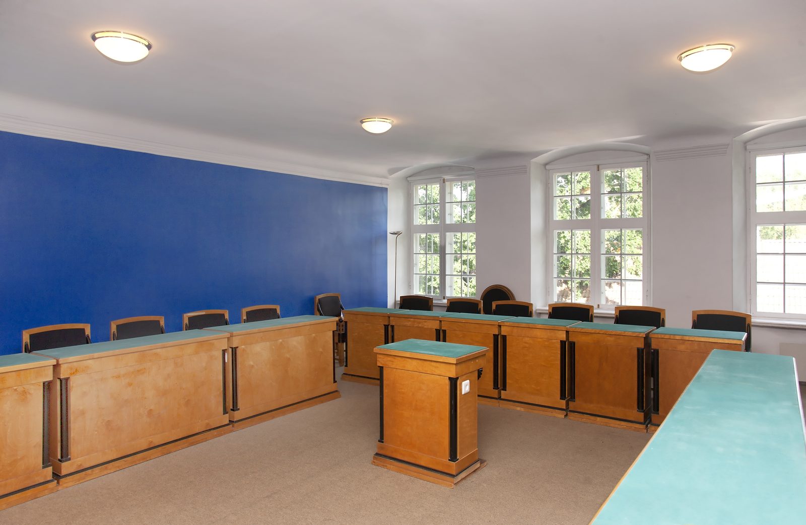 Empty public meeting chamber with rows of wooden desks and chairs, a central podium, a blue wall and tall windows letting in daylight
