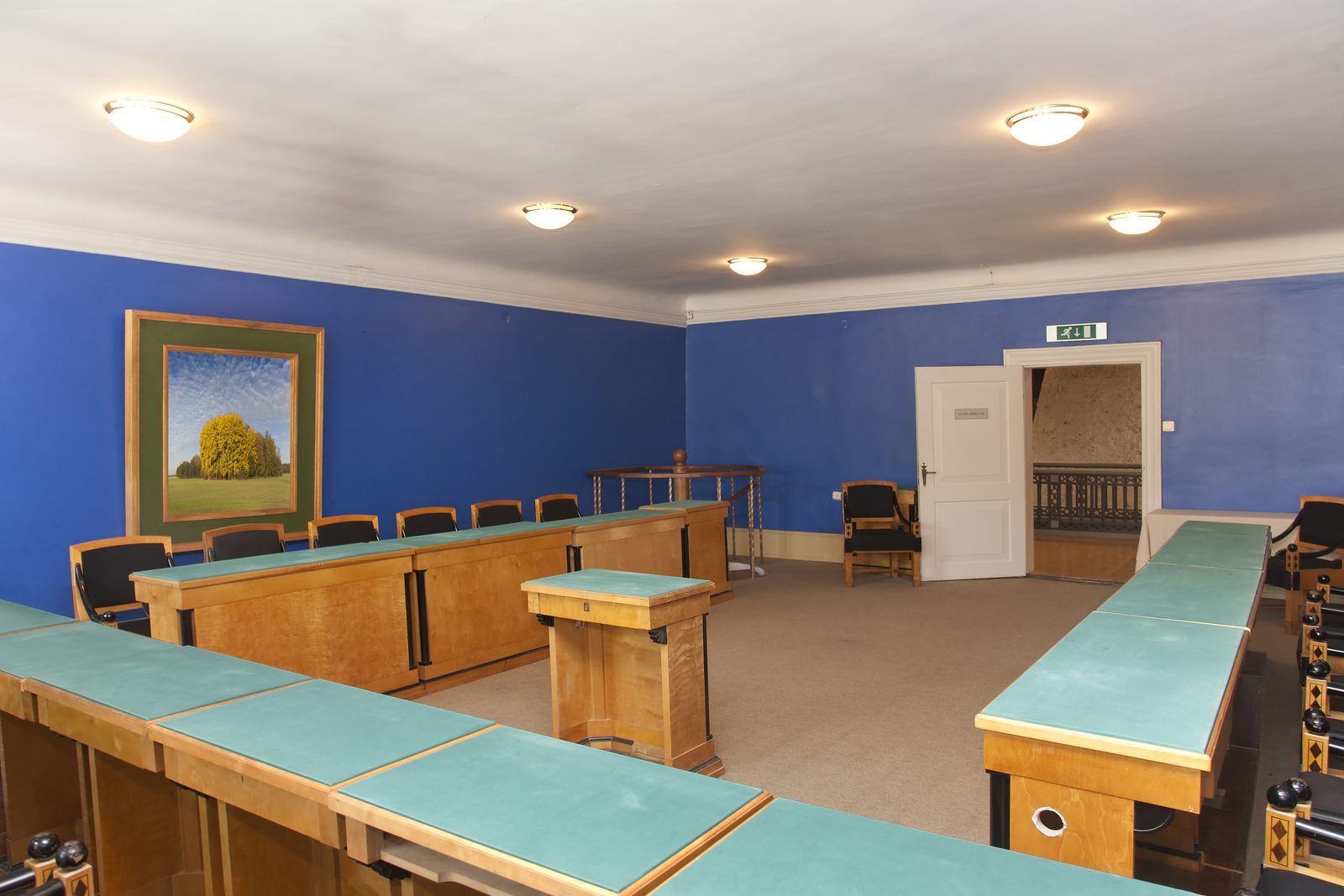 Empty municipal meeting room with rows of wooden desks, a central podium and blue walls under ceiling lights