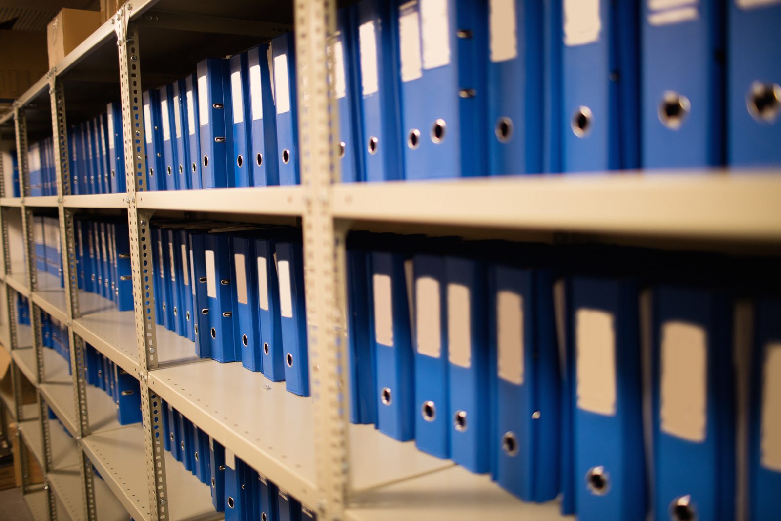 Rows of blue lever arch binders with blank spine labels fill metal storage shelves in a records room