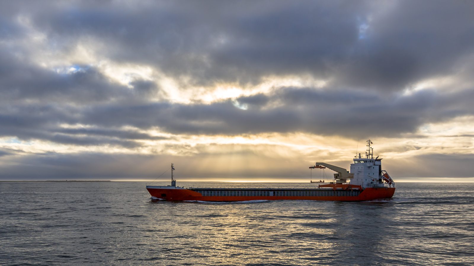 A red-hulled freight ship moves across open gray water beneath heavy clouds, with low sunlight breaking through near the horizon.
