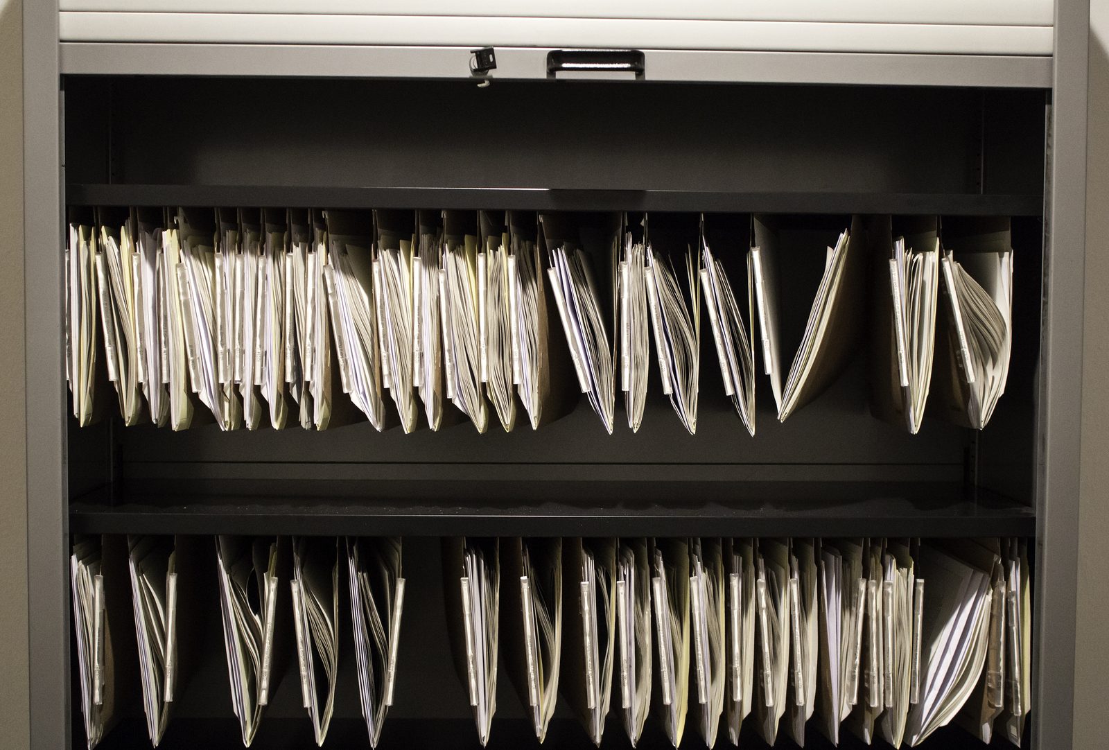 Rows of white paper files hang in a gray office filing cabinet with its roll-up door open, lit from above.