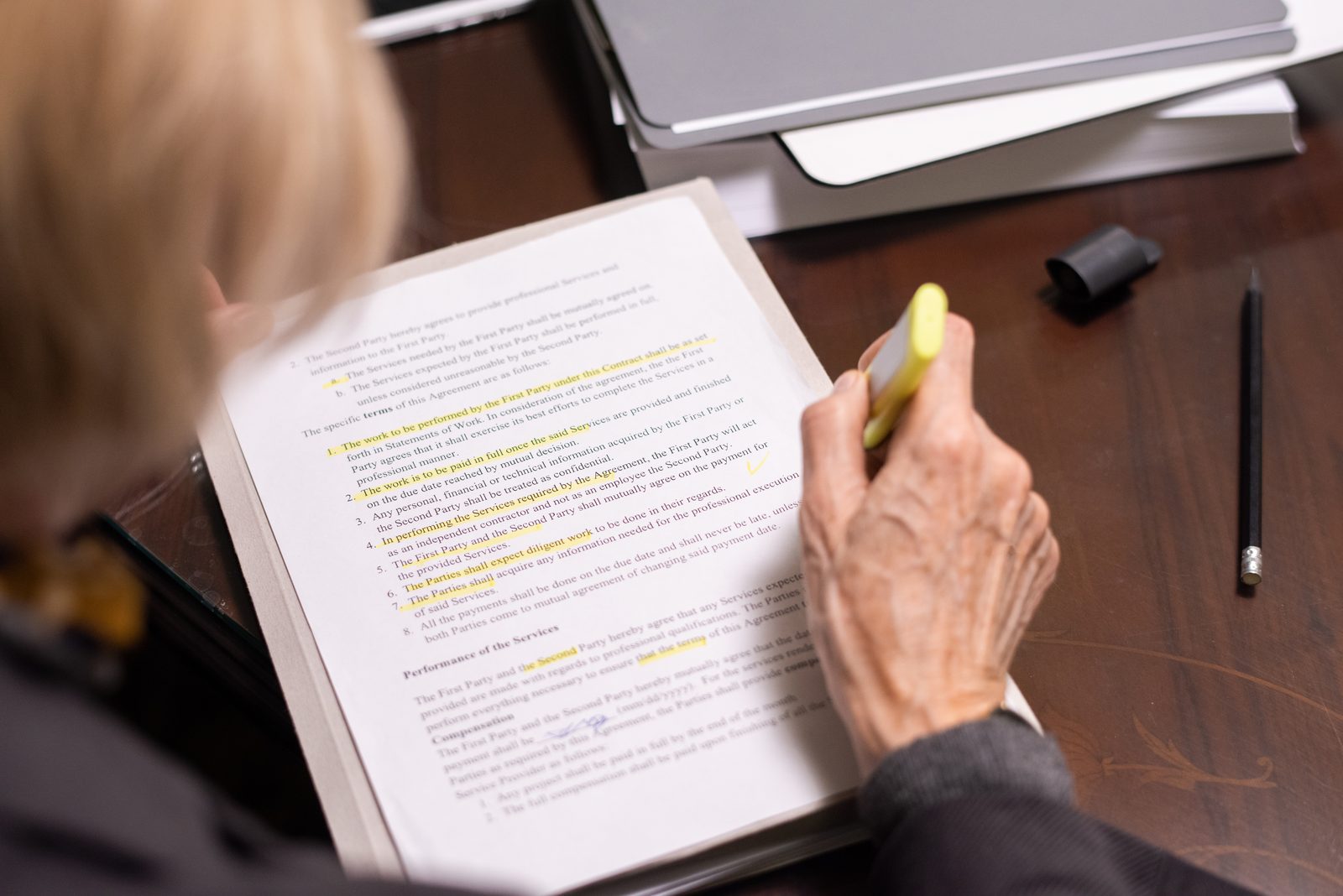 A hand holds a yellow highlighter over a printed document with marked lines, a pen and folders resting on a wooden desk.