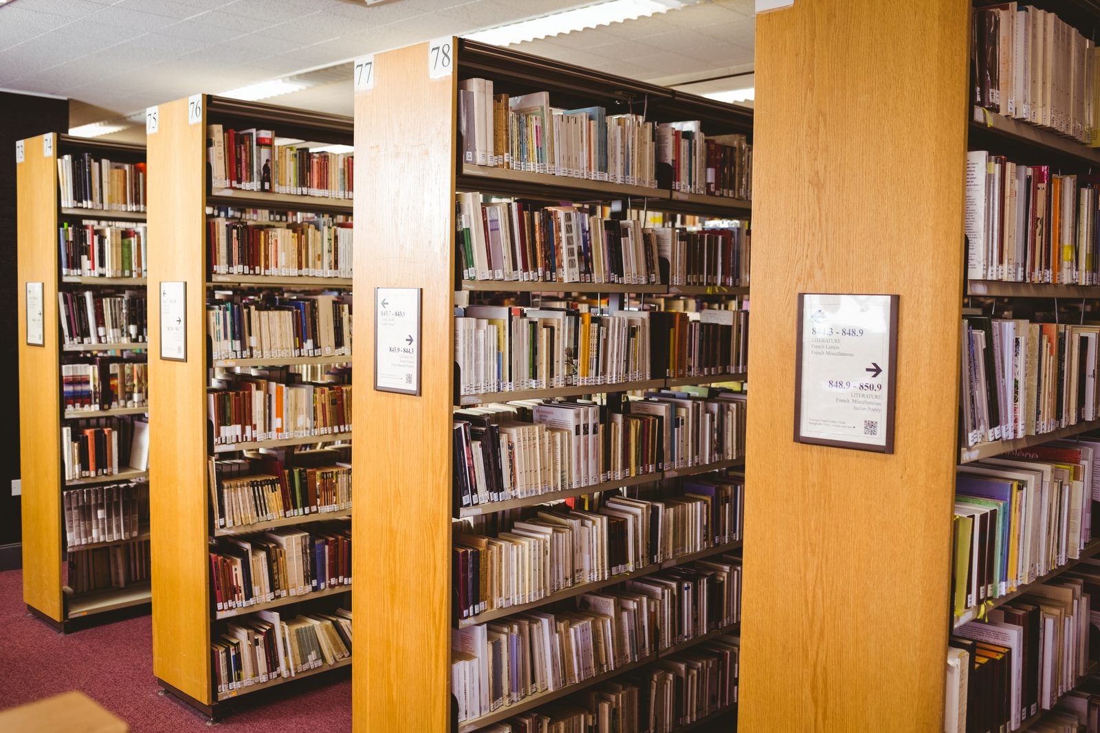 Tall wooden library shelving units stand in rows, filled with bound volumes and marked with numbered end panel signs