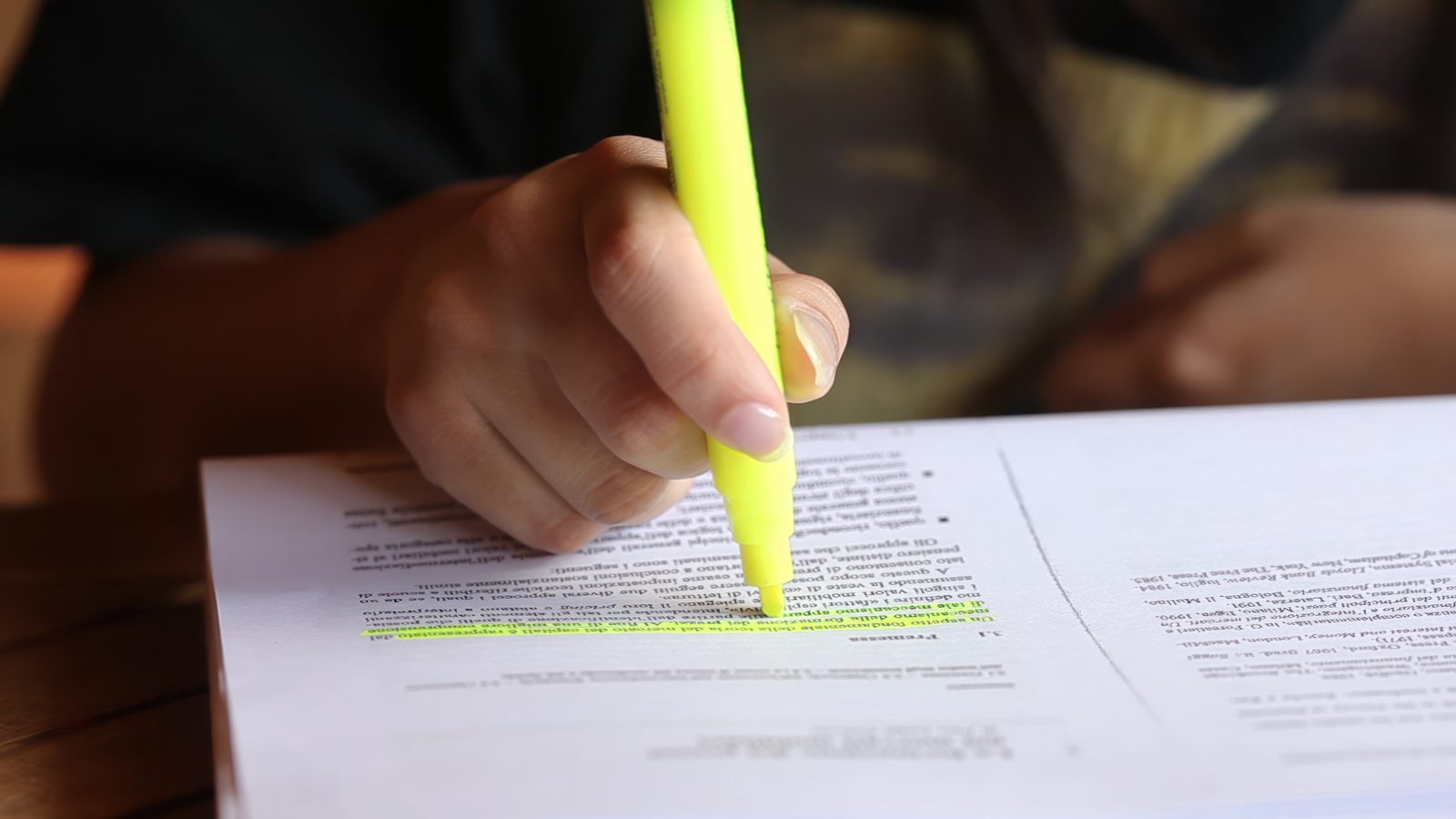 A hand draws a yellow highlighter across a line of text on a printed multi-page document resting on a wooden table