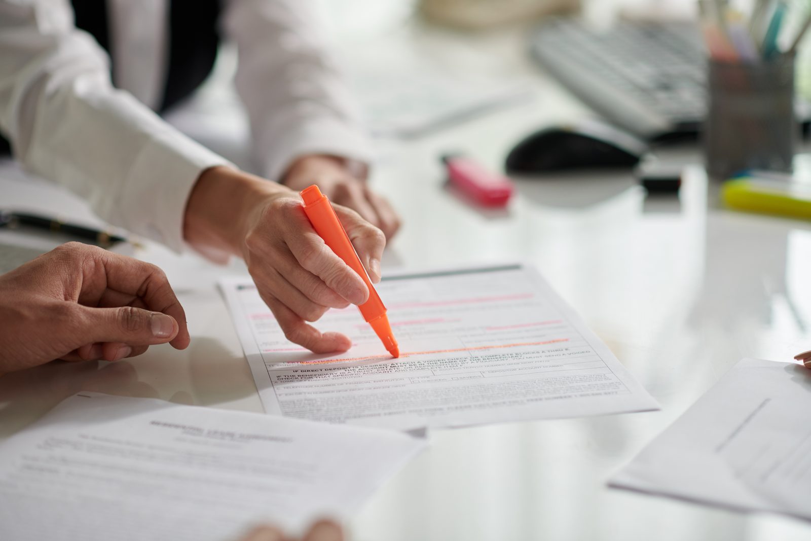 Two people at a white office desk review a printed document while one marks lines of text with an orange highlighter.