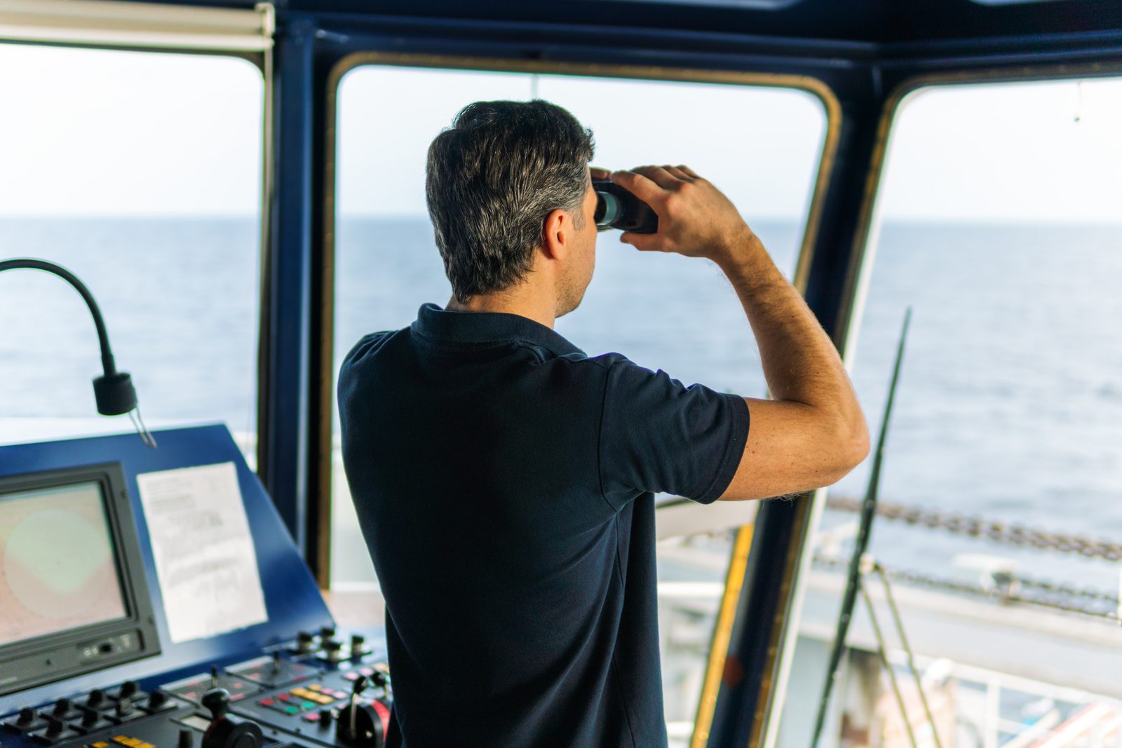 A ship officer on the bridge raises binoculars toward open sea, standing beside a navigation console with a monitor and control panel.