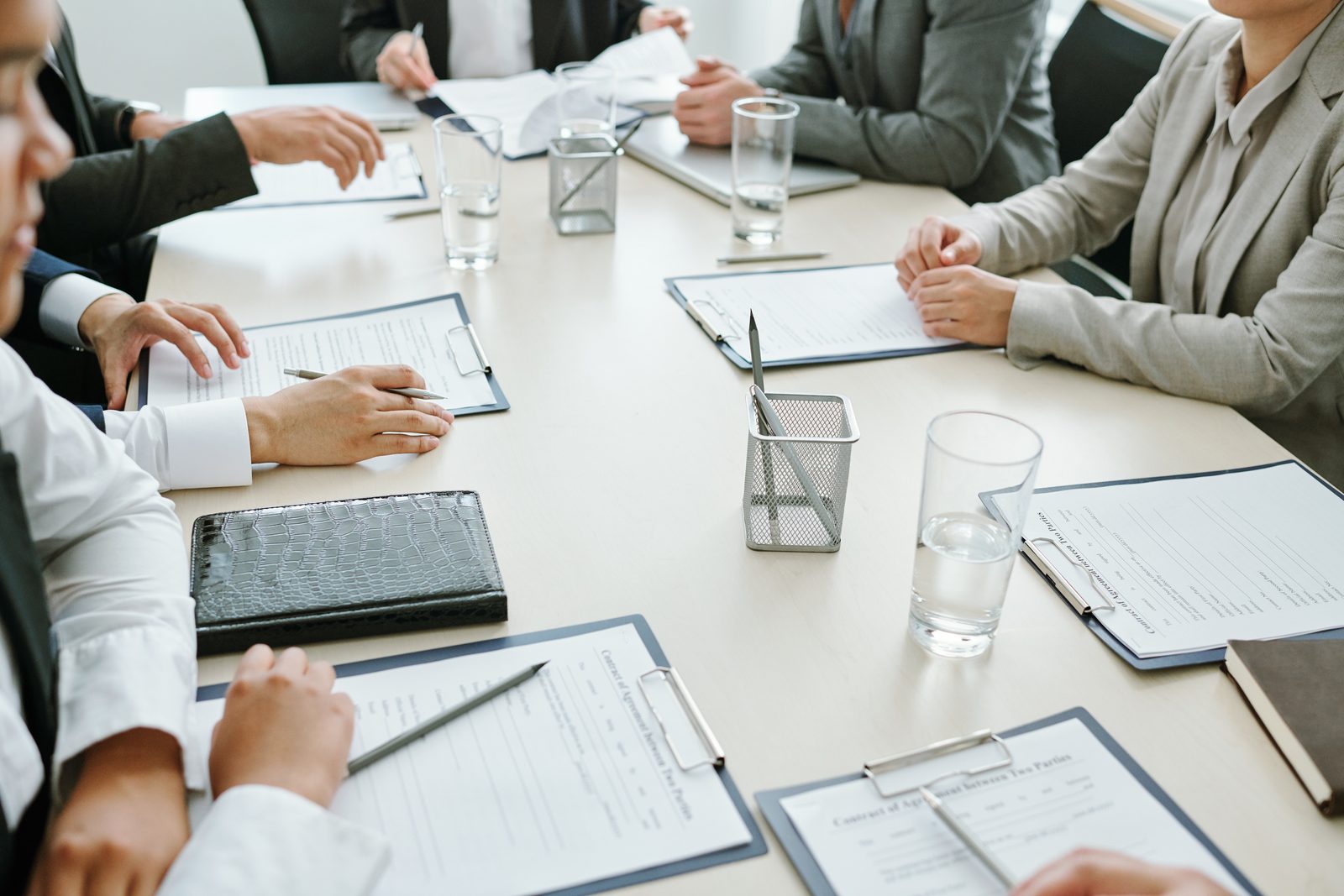 People seated around a pale meeting table read printed pages on clipboards, with pens, water glasses and a pencil cup between them.