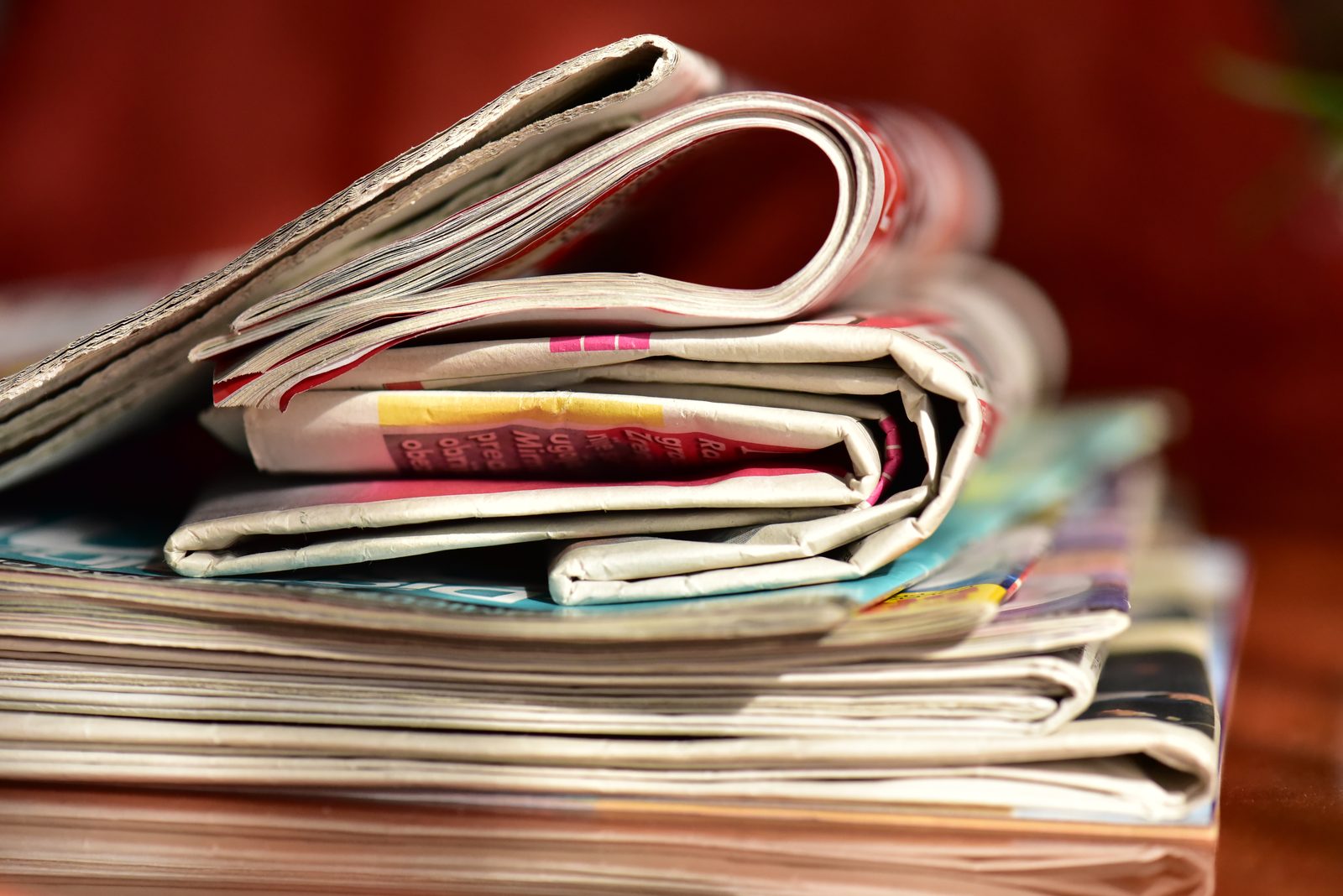 A stack of folded newspapers and magazines rests on a wooden table against a blurred red background.