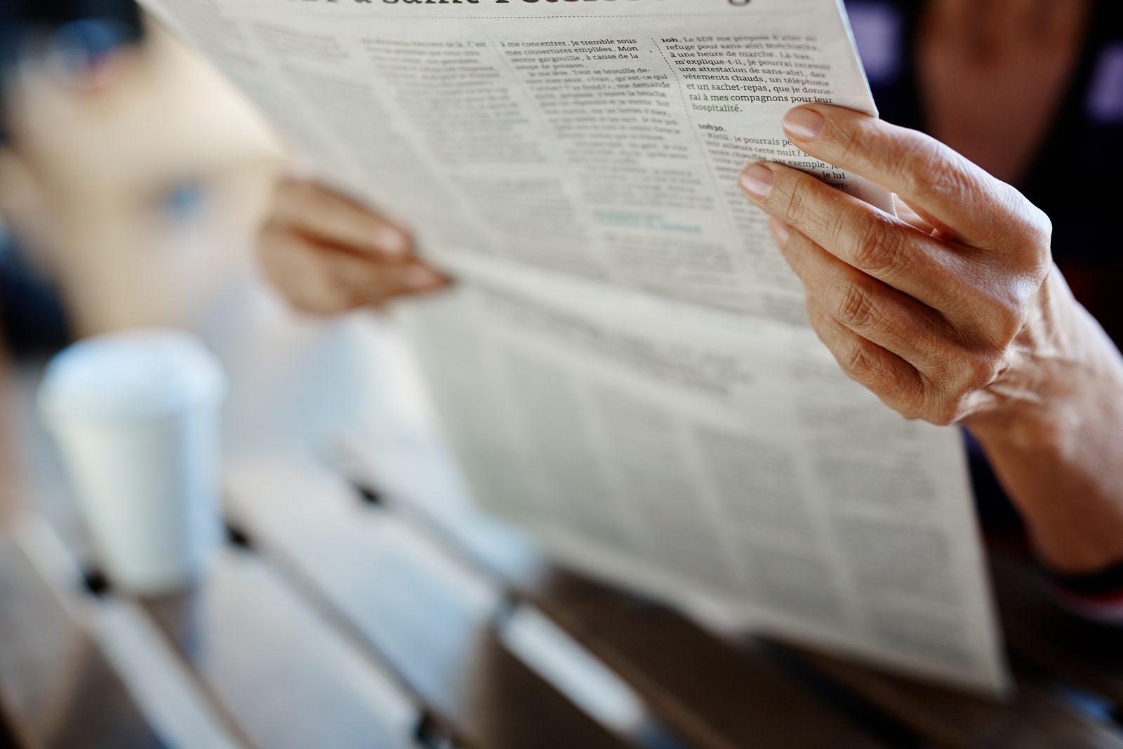 A reader holds an open broadsheet newspaper in both hands, its columns of small print in focus above a blurred cafe table