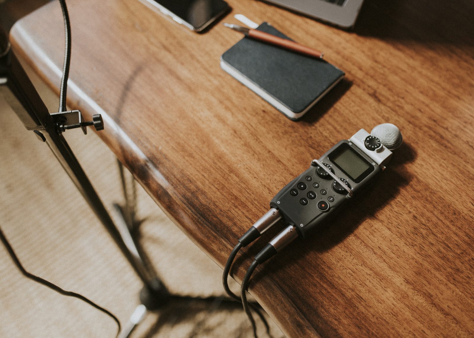 A handheld audio recorder with cables rests on a wooden table beside a closed black notebook, a pen and a microphone stand.