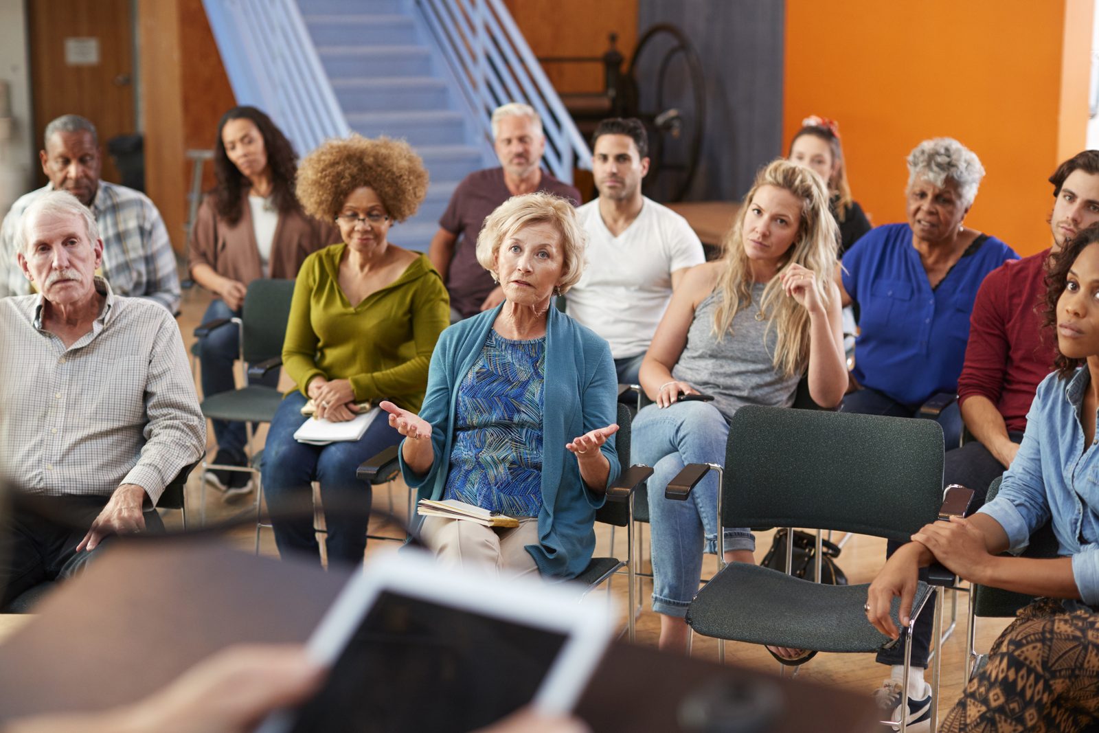 Residents seated in rows at a community center meeting as a woman in a teal cardigan speaks with open hands