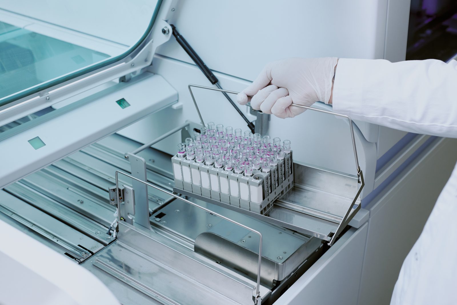 Gloved laboratory worker lowering a rack of capped sample tubes into an automated analyzer in a bright clinical lab