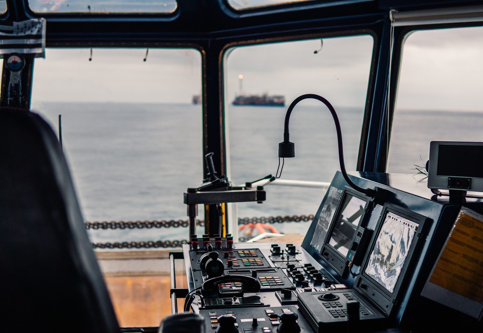A ship's bridge console with radar screens, radio handset and control levers faces windows onto a gray sea and a distant vessel.