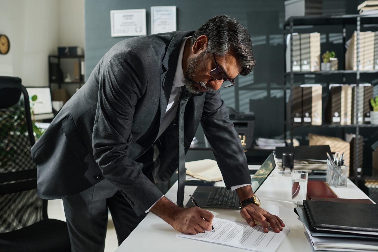 Man in a dark suit and glasses leaning over an office desk to sign a printed document beside a laptop and binders