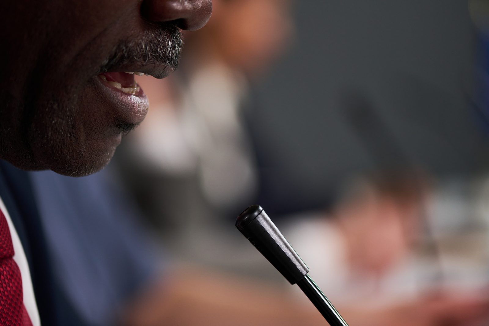 Man in a suit and red tie speaks into a desk microphone during a meeting, other attendees blurred behind him