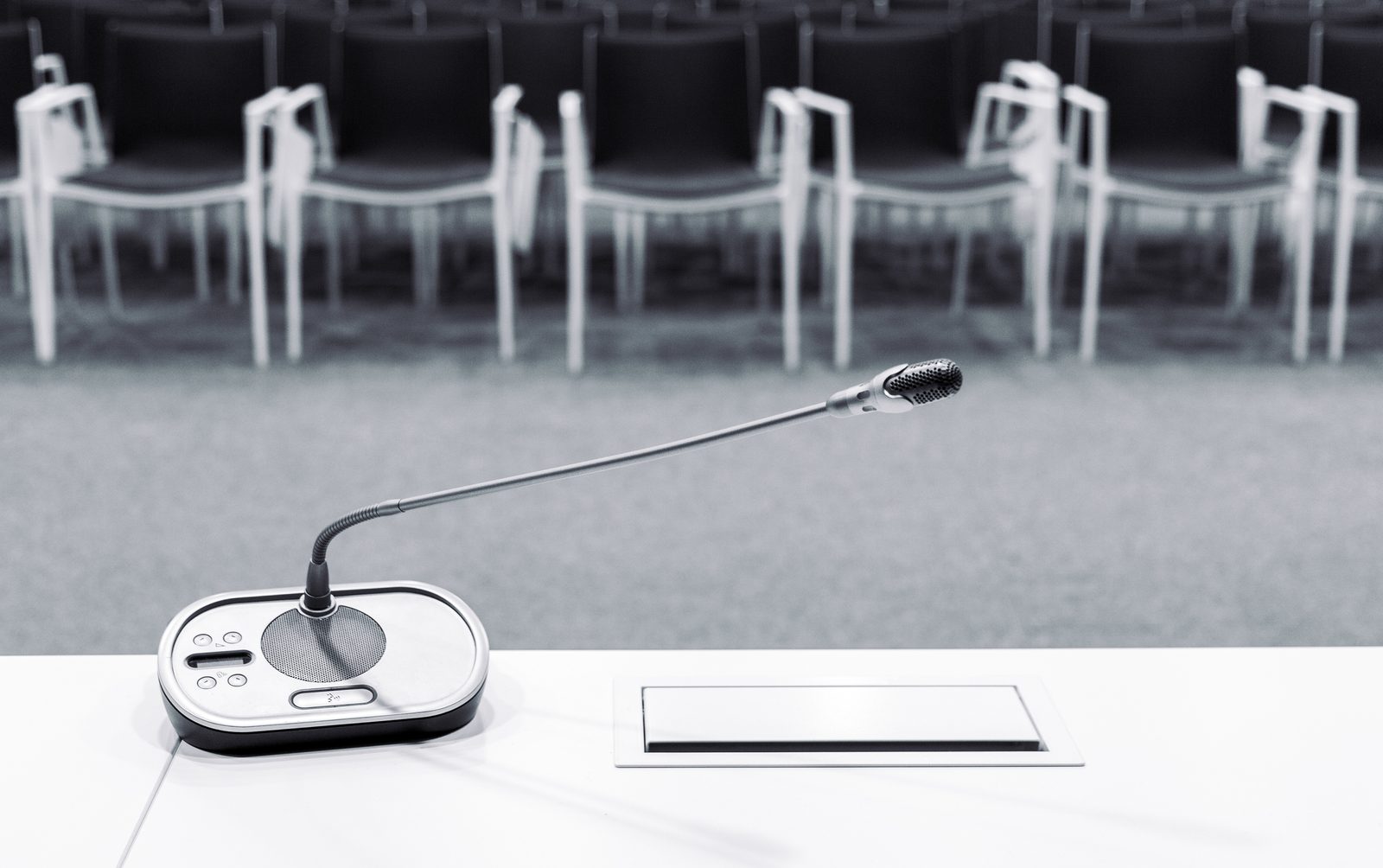 A conference microphone on an empty white table faces rows of vacant chairs in a black and white meeting room.